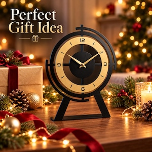 May include: A black and gold table clock with a circular frame and a central dial featuring the numbers 3, 6, 9, and 12. The clock has thin gold hands and is positioned on a flat base. The text "Perfect Gift Idea" is visible in the upper left corner.