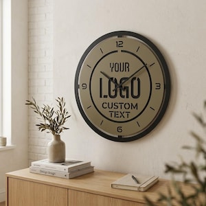 May include: A round wall clock with a black frame and a tan face. The clock features black hour and minute hands, black numerals at the 3, 6, 9, and 12 positions, and the black text YOUR LOGO CUSTOM TEXT printed in the center of the clock face.