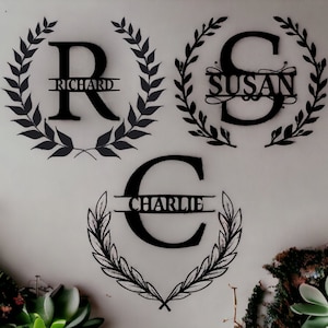 K&ouml;nnte beinhalten: Drei schwarze Wanddekorationen aus Metall mit einem Kranzdesign und einem Buchstaben in der Mitte. Die Buchstaben sind "R", "S" und "C". Die Namen "Richard", "Susan" und "Charlie" sind auf den Kr&auml;nzen geschrieben.