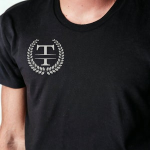 K&ouml;nnte beinhalten: Ein schwarzes T-Shirt mit einem silbernen Monogramm-Design, das den Buchstaben "T" zeigt, umgeben von einem Lorbeerkranz.