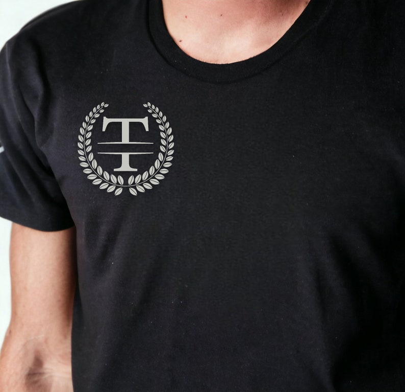 K&ouml;nnte beinhalten: Ein schwarzes T-Shirt mit einem silbernen Monogramm-Design, das den Buchstaben "T" zeigt, umgeben von einem Lorbeerkranz.