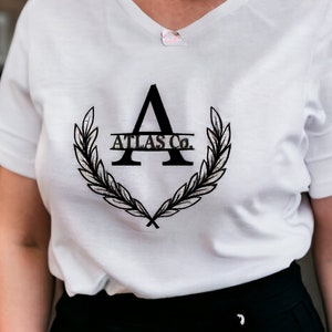 K&ouml;nnte beinhalten: Wei&szlig;es T-Shirt mit einer schwarzen Grafik eines stilisierten Buchstabens A mit einem Lorbeerkranz und dem Text "ATLAS Co."