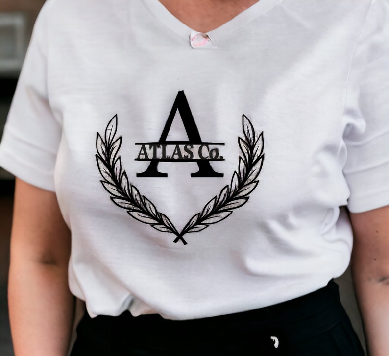 K&ouml;nnte beinhalten: Wei&szlig;es T-Shirt mit einer schwarzen Grafik eines stilisierten Buchstabens A mit einem Lorbeerkranz und dem Text "ATLAS Co."