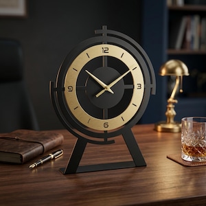 May include: A table clock featuring a matte black circular frame with a gold-toned inner ring. The clock face displays gold-colored hands and numerals at the 12, 3, 6, and 9 o'clock positions, supported by a sturdy black metal stand base.