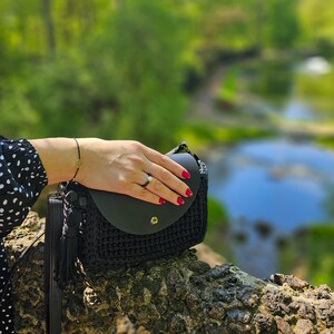 Puede incluir: Un bolso de mano de crochet negro con una solapa de cuero negro y un borla. El bolso está sobre un tronco de árbol.