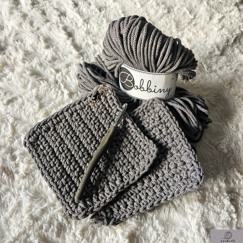 May include: Two gray crocheted squares with a gray crochet hook and a ball of gray yarn with a white label that says "Bobbiny".