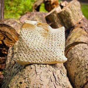 Op de afbeelding: Een beige gehaakte handtas met een herhalend stervormig patroon over het hele lichaam. De tas heeft een gestructureerde rechthoekige vorm met een stevig geweven bovenrand en geïntegreerde handvatten, met een gedetailleerde textuur.