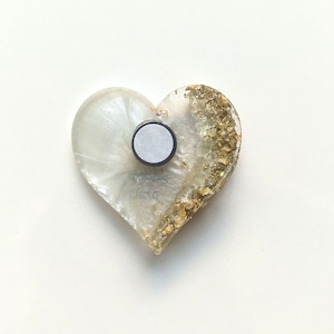 May include: Heart-shaped magnet with a pearlescent white resin base and gold leaf accents. The magnet has a silver-colored backing and the text "H2TO" is visible. A decorative item for the home.