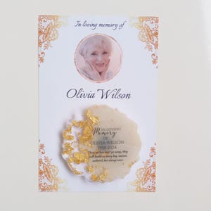 May include: A white memorial card with a gold border and a photo of a woman. The card reads "In loving memory of Olivia Wilson 1958-2024. These we love don't go away, they walk beside us every day, unseen, unheard, but always near."