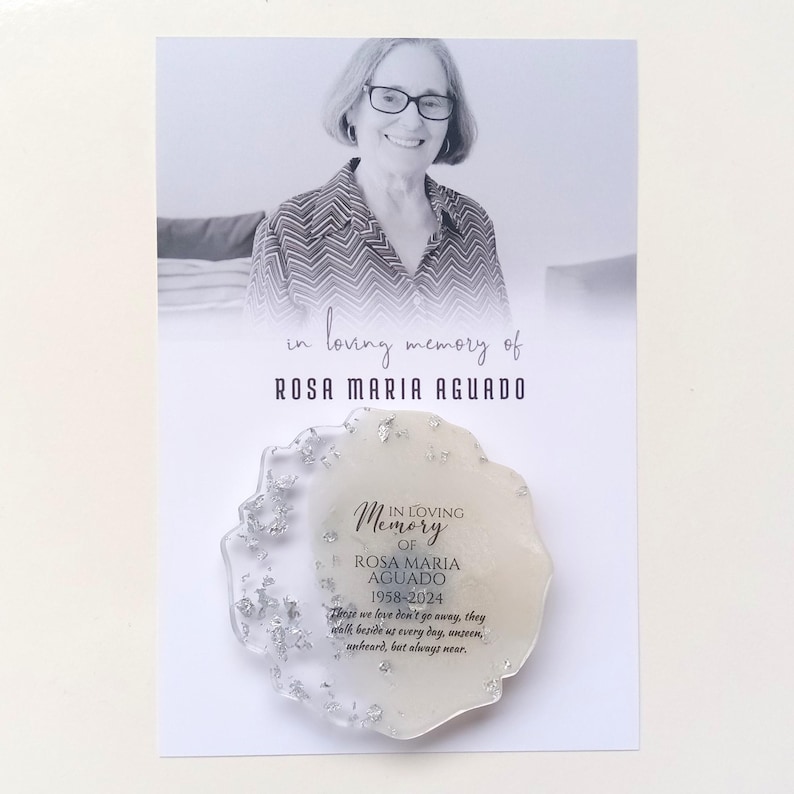 May include: A white memorial plaque with silver glitter embedded in the resin. The plaque has a white background with black text that reads "In Loving Memory of Rosa Maria Aguado 1958-2024". The text continues with a quote: "Those we love don't go away, they walk beside us every day, unseen, unheard, but always near."