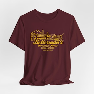 Könnte beinhalten: Ein kastanienbraunes Kurzarm-T-Shirt mit einer gelben Strichzeichnung eines großen Gebäudes. Der Text lautet Kellerman's Mountain House, Catskills, New York, und I HAD THE TIME OF MY LIFE in einer stilisierten Schriftart auf der Brust.