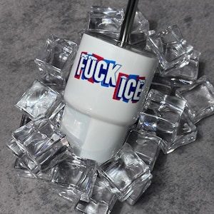 May include: A white tumbler with the words "FUCK ICE" in red and blue lettering, surrounded by clear ice cubes. The tumbler has a silver straw and a white base. The background is a gray surface.