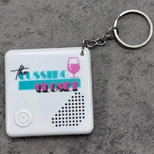 May include: White square keychain with the text "The Cussing Closet" in black and pink, with a teal stripe and a pink wine glass graphic. The keychain has a silver keyring and a small button on the front.