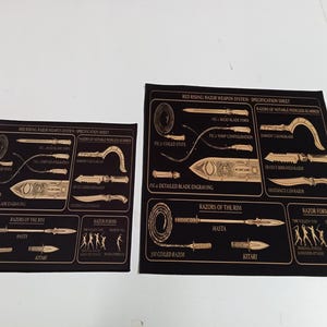May include: Two black rectangular prints featuring gold line art diagrams titled Red Rising: Razor Weapon System - Specification Sheet. The graphics display various blade forms, coiled states, detailed engravings, and weapon forms with labels.