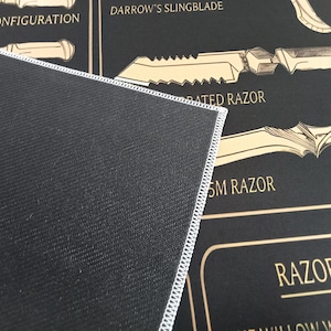 May include: A black rectangular mat with a white stitched edge, featuring a graphic print of various bladed weapons in tan and gold. The text includes labels such as WHIP CONFIGURATION, DARROW'S SLINGBLADE, SERRATED RAZOR, SM RAZOR, and RAZOR FORM.