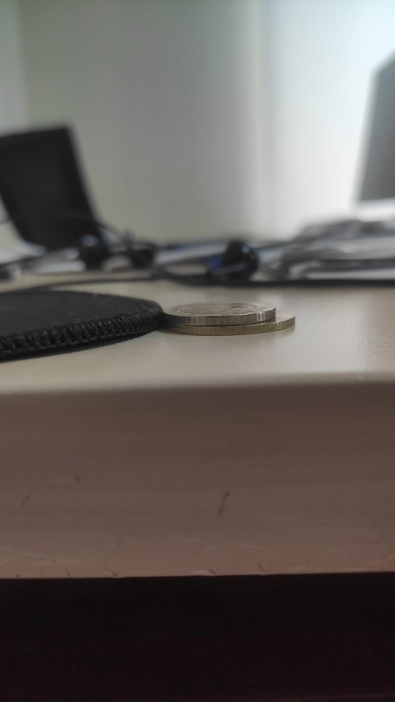 May include: Two circular metallic coins stacked on top of each other. The top coin is silver in color with a smooth edge, while the bottom coin is gold in color with a reeded edge. The coins are positioned horizontally on a flat surface.