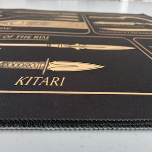 May include: A black rectangular desk mat featuring gold-colored line art illustrations of various bladed weapons. The text includes labels such as Razors of the Rim, Hasta, and Kitari. The mat has a stitched edge and a smooth, flat surface texture.