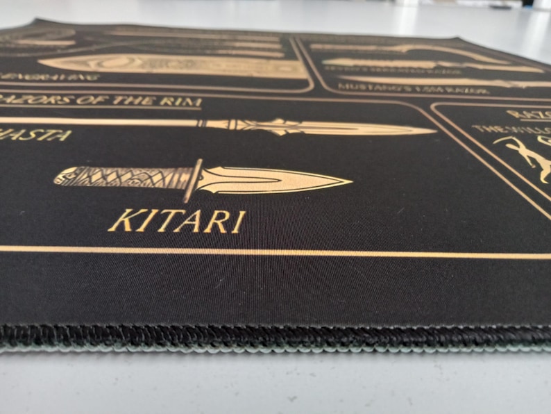 May include: A black rectangular desk mat featuring gold-colored line art illustrations of various bladed weapons. The text includes labels such as Razors of the Rim, Hasta, and Kitari. The mat has a stitched edge and a smooth, flat surface texture.