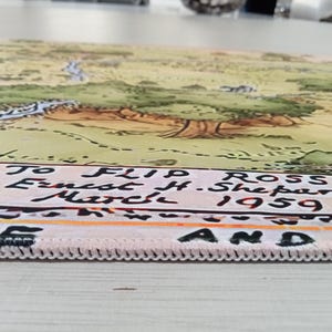 May include: A rectangular fabric mat featuring a printed illustration of a landscape with trees and a river. The bottom edge displays the text: To Flip Ross Ernest H. Shepard March 1959. The mat has a white stitched border along the bottom edge.