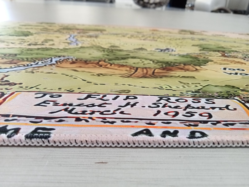 May include: A rectangular fabric mat featuring a printed illustration of a landscape with trees and a river. The bottom edge displays the text: To Flip Ross Ernest H. Shepard March 1959. The mat has a white stitched border along the bottom edge.