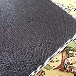 May include: A rectangular desk mat featuring a textured black rubber underside and a white stitched edge. The top surface displays a portion of a colorful illustrated map with visible land features and the partial text OV and HO.