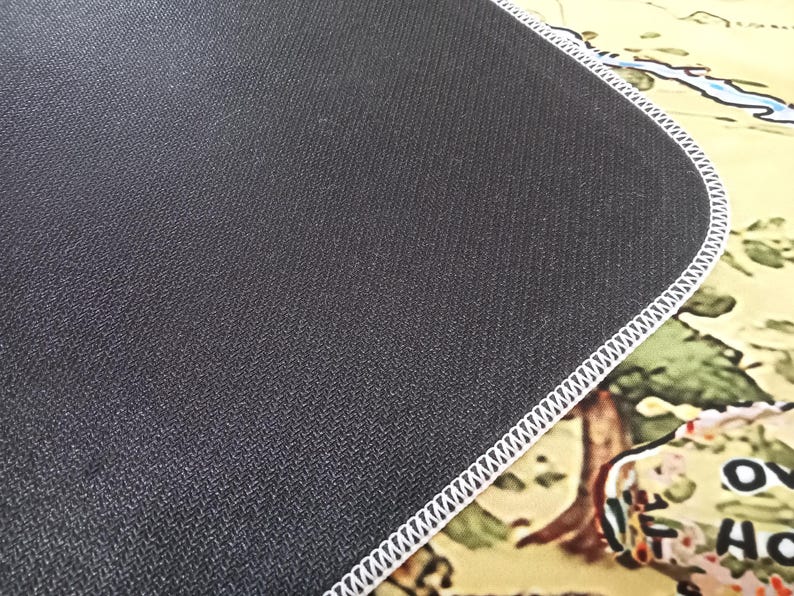 May include: A rectangular desk mat featuring a textured black rubber underside and a white stitched edge. The top surface displays a portion of a colorful illustrated map with visible land features and the partial text OV and HO.