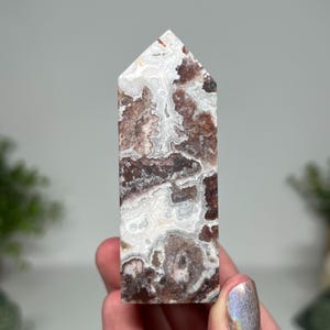 May include: A tall, rectangular crystal point with a flat top. The stone features a swirling pattern of white, brown, and reddish-brown colors. The crystal is held in a hand, showcasing its polished surface and natural variations.