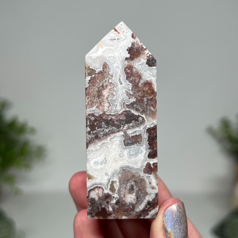 May include: A tall, rectangular crystal point with a flat top. The stone features a swirling pattern of white, brown, and reddish-brown colors. The crystal is held in a hand, showcasing its polished surface and natural variations.
