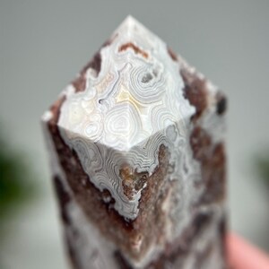 May include: A close-up of a polished agate crystal tower. The stone features a mix of brown, white, and cream colors with intricate banded patterns. The top is a pointed, faceted shape.