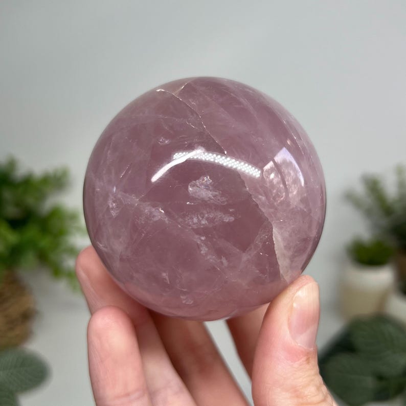 May include: A polished, spherical rose quartz crystal ball. The sphere is a soft pink color with natural variations in tone and texture. The crystal is held in a hand, with a blurred background of greenery.