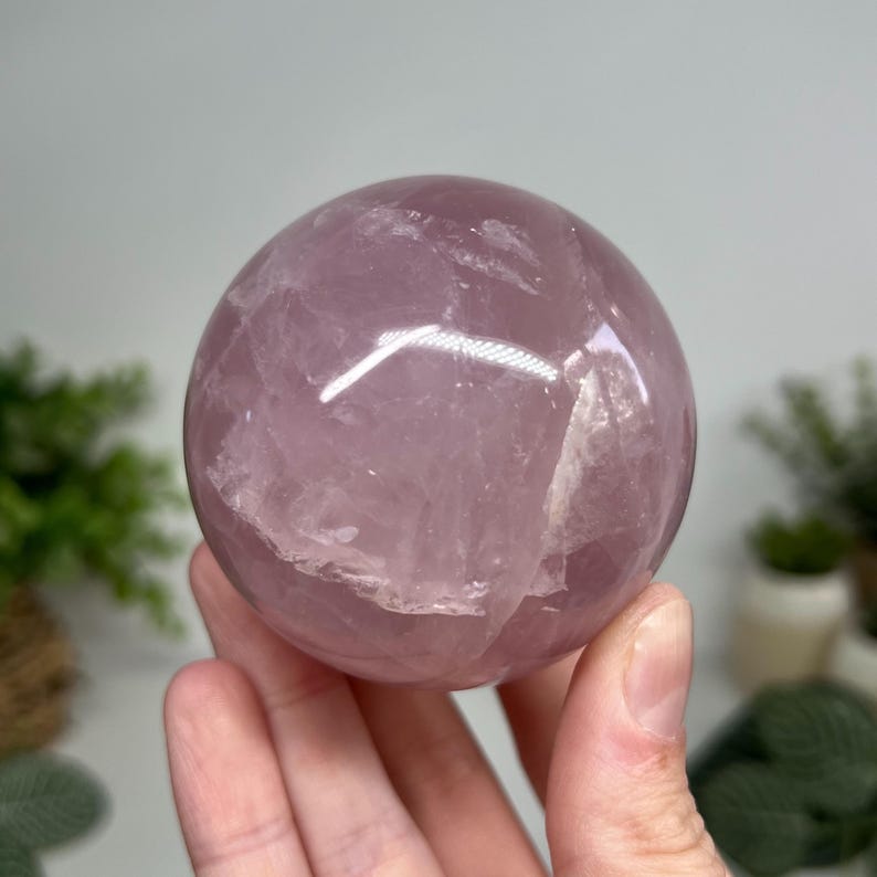May include: A polished, spherical rose quartz crystal ball. The sphere is a soft pink color with internal crystalline structures visible. The surface reflects light, creating highlights. The crystal is held in a hand, with a blurred background of greenery.
