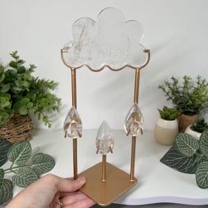 May include: A decorative stand featuring a clear, cloud-shaped crystal top and three clear, teardrop-shaped crystal pendants suspended from a gold-toned metal frame. The structure includes a square gold base and two vertical metal rods.