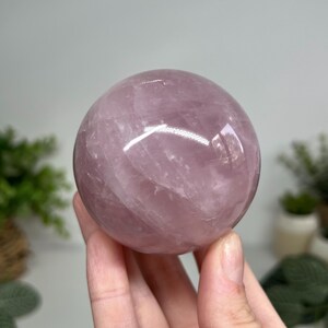 May include: A polished, translucent rose quartz sphere. The sphere is a soft pink color with subtle internal veining. The smooth surface reflects light, creating highlights. The sphere is held in a hand, with a blurred background.