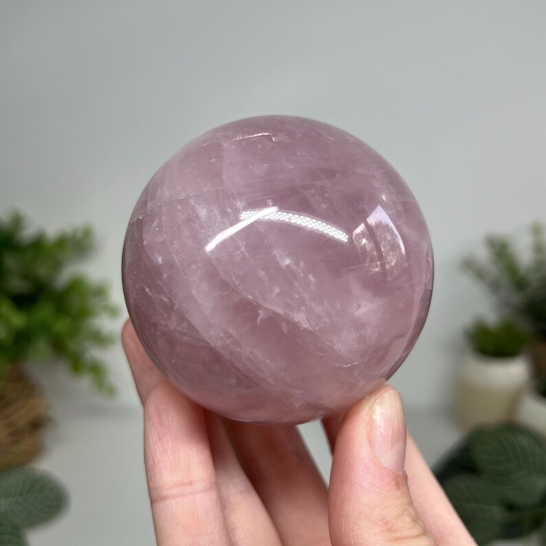 May include: A polished, translucent rose quartz sphere. The sphere is a soft pink color with subtle internal veining. The smooth surface reflects light, creating highlights. The sphere is held in a hand, with a blurred background.