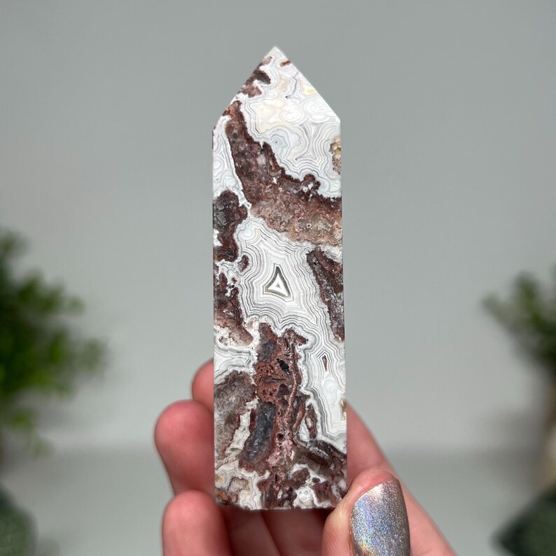 May include: A tall, rectangular crystal point with a pointed top. The stone features a mix of white, brown, and gray patterns. The crystal is held in a hand, showcasing its polished surface and natural variations. The crystal is a decorative item.