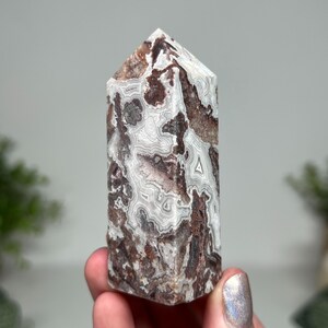 May include: A polished agate crystal tower with a pointed top. The stone features a mix of white, brown, and gray patterns. The crystal is held in a hand, showcasing its natural beauty and unique veining.
