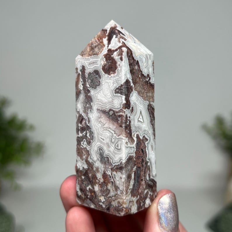 May include: A polished agate crystal tower with a pointed top. The stone features a mix of white, brown, and gray patterns. The crystal is held in a hand, showcasing its natural beauty and unique veining.