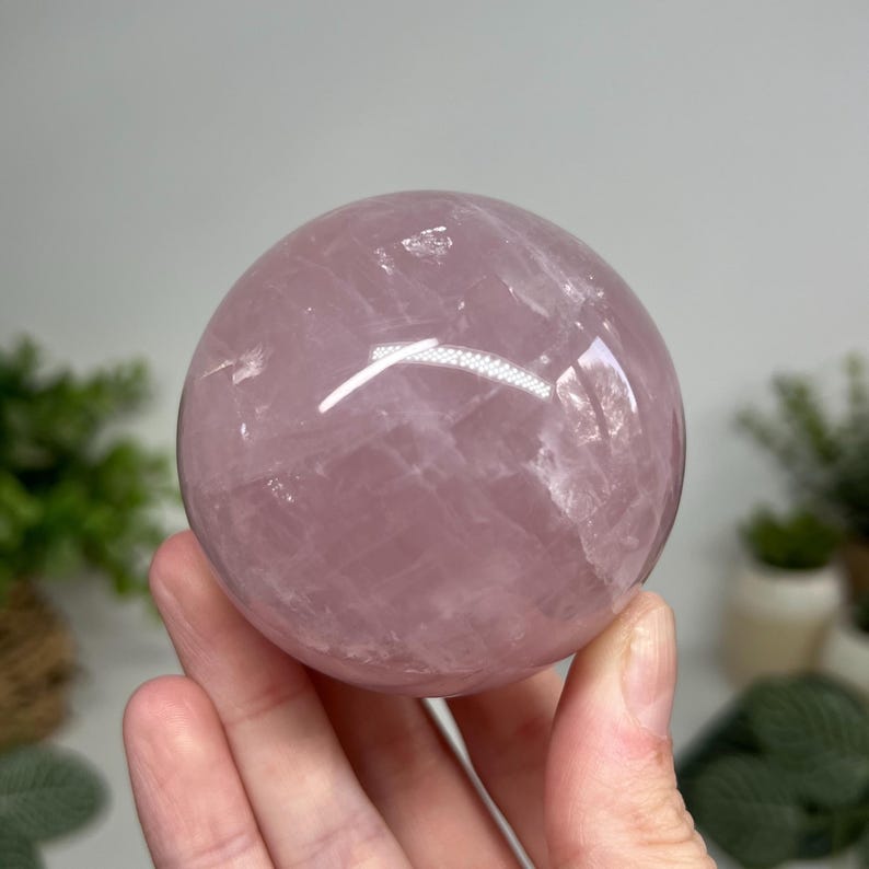 May include: A polished rose quartz sphere, showcasing a soft pink hue. The translucent stone has natural veining and a smooth, rounded shape. The sphere is held in a hand, with a blurred background.
