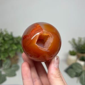 May include: A polished, spherical carnelian stone featuring a deep orange-red hue and a central, irregular cavity filled with small, sparkling, light-coloured crystals. The surface of the sphere is smooth and glossy, showcasing natural banding.