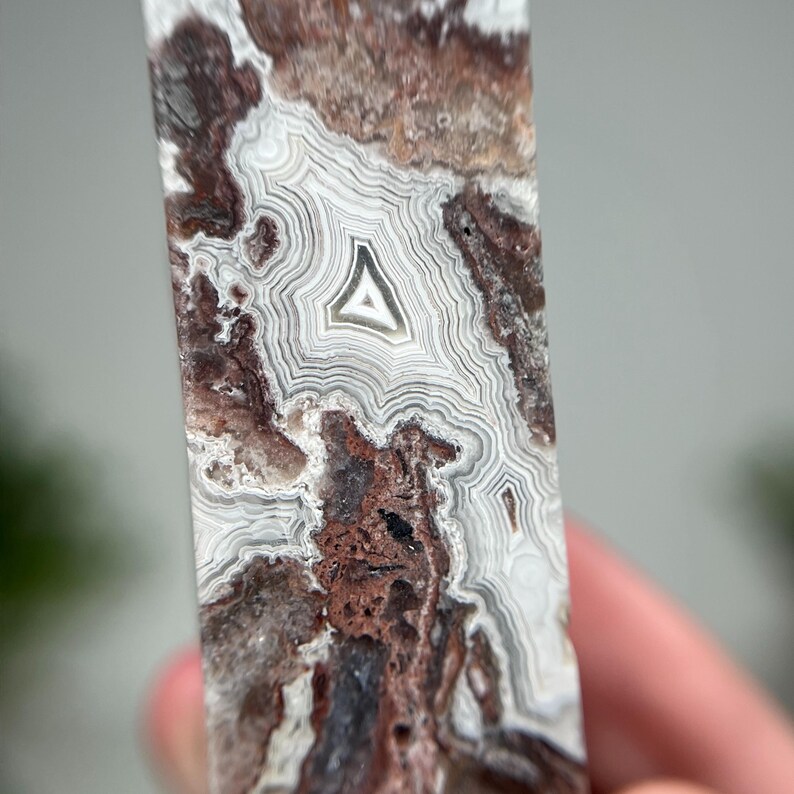 May include: A close-up of a polished stone with a unique pattern. The stone features concentric bands of white, brown, and reddish-brown hues. A central triangle shape is visible within the stone's intricate design. The stone's surface is smooth and reflective.