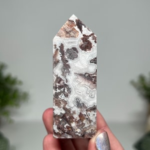 May include: A polished, rectangular crystal tower with a pointed top. The stone features a complex pattern of swirling white, brown, and gray bands. The crystal is held in a hand, with a blurred background.