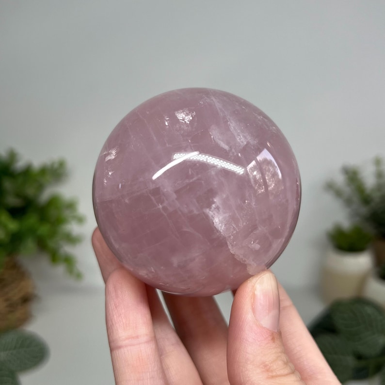 May include: A polished rose quartz sphere, showcasing a soft pink hue with natural veining. The sphere is held in a hand, with a bright reflection on its surface. The crystal ball is a decorative item.