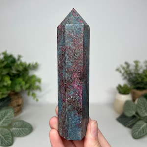 May include: A polished hexagonal crystal tower featuring a mottled blend of deep magenta, dark red, and slate blue mineral inclusions throughout its vertical structure, terminating in a sharp, multi-faceted point at the top.