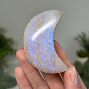 May include: A crescent moon-shaped polished gemstone with a translucent, off-white base and a vibrant, iridescent blue flash across its surface. The stone features a smooth, curved edge and a flat, textured interior, displaying natural mineral inclusions.