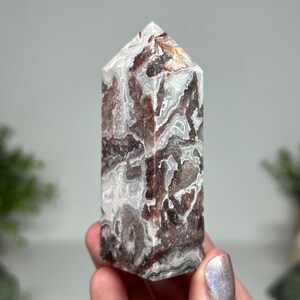 May include: A polished, rectangular crystal tower with a pointed top. The stone features a swirling pattern of white, brown, and gray. The crystal is held in a hand, with a blurred background.