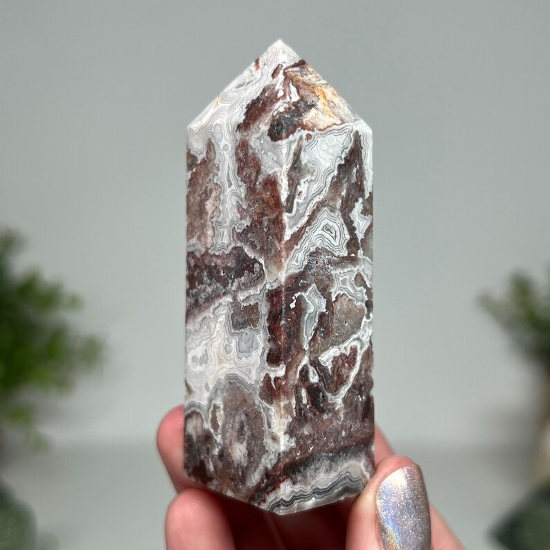 May include: A polished, rectangular crystal tower with a pointed top. The stone features a swirling pattern of white, brown, and gray. The crystal is held in a hand, with a blurred background.