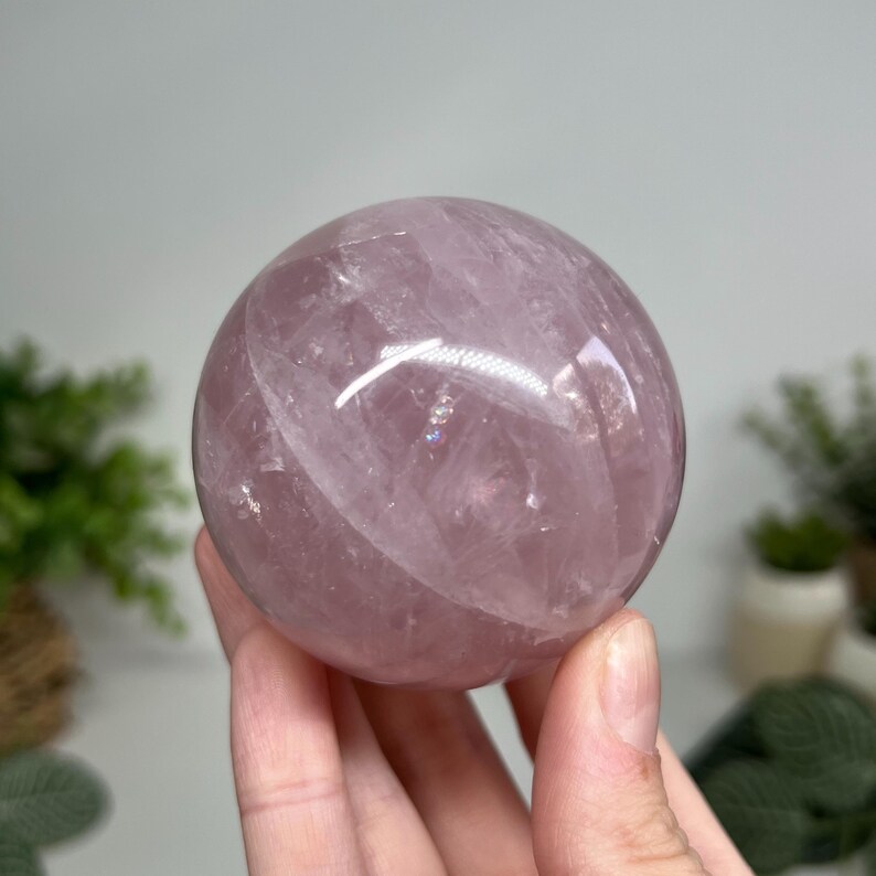May include: A translucent, light purple crystal sphere. The polished gemstone is held in a hand, showcasing its smooth, rounded shape and internal patterns. The sphere reflects light, highlighting its color and clarity.