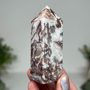 May include: A polished, rectangular crystal tower with a pointed top. The stone features a complex pattern of swirling white, brown, and gray bands. The crystal is held in a hand, with a blurred background.