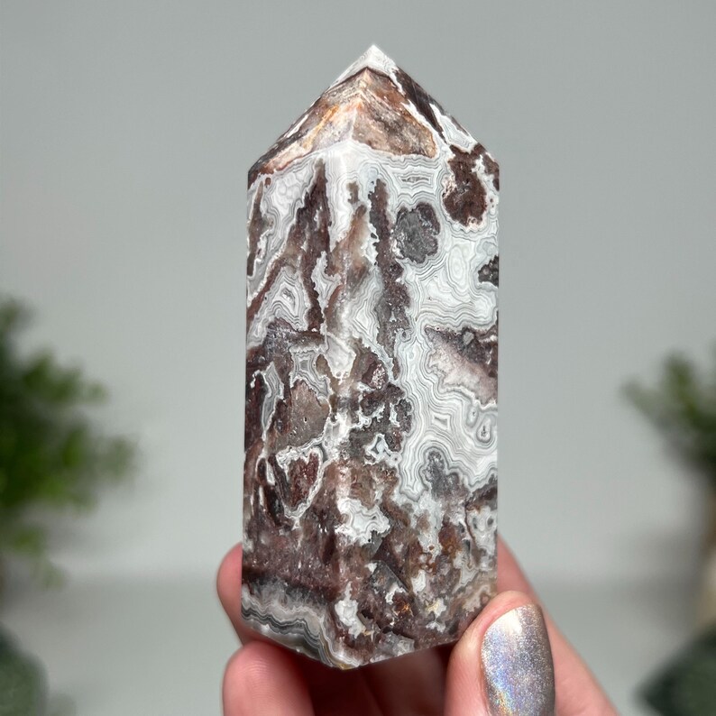 May include: A polished, rectangular crystal tower with a pointed top. The stone features a complex pattern of swirling white, brown, and gray bands. The crystal is held in a hand, with a blurred background.