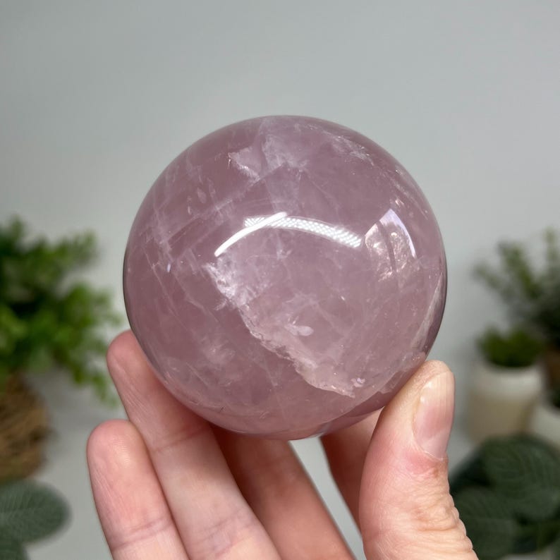 May include: A polished, spherical rose quartz crystal ball. The sphere is a soft pink color with subtle variations in tone and internal veining. The surface reflects light, creating highlights and shadows. The crystal is held in a hand.